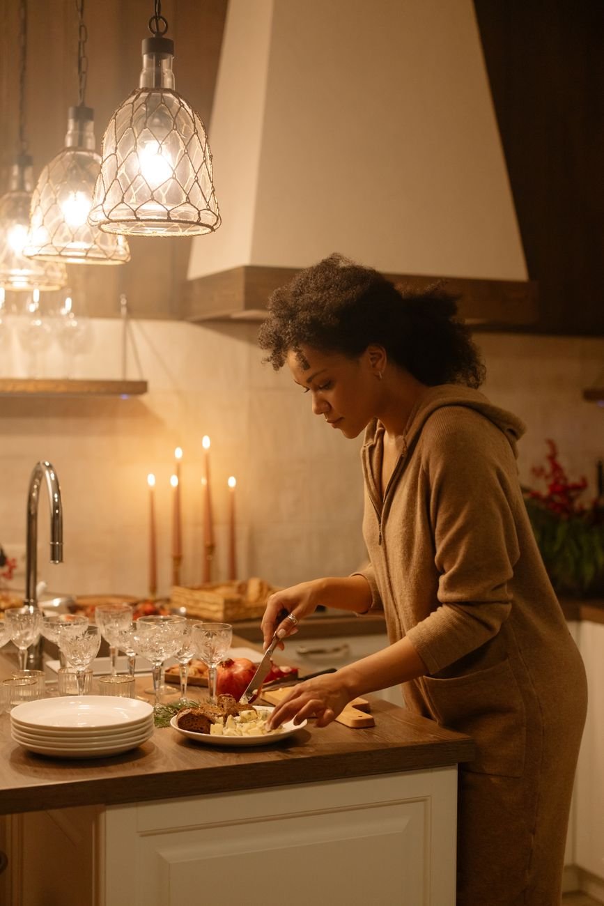 A woman with curly hair prepares a meal in a warmly lit kitchen, surrounded by candles and tableware.