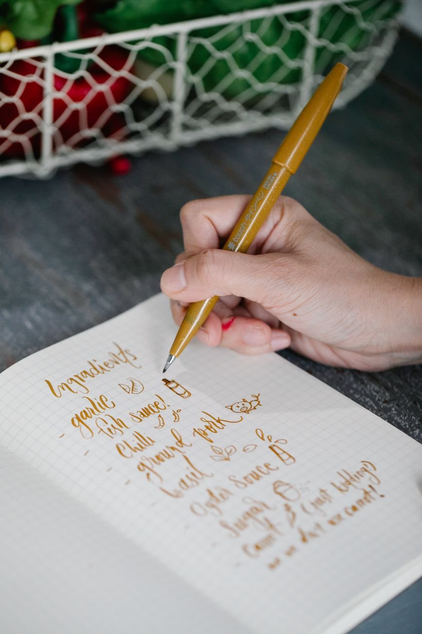 Close-up of a hand writing ingredients list with a golden pen on a notepad.